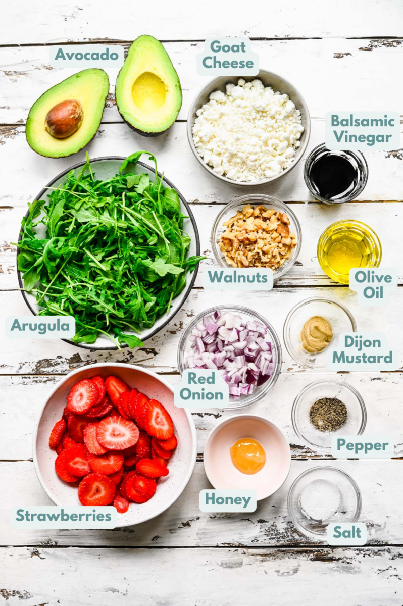 Ingredients for strawberry arugula salad in white bowls: arugula, strawberries, avocado, red onion, walnuts, goat cheese, olive oil, balsamic vinegar, dijon mustard, honey, salt, pepper