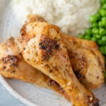 3 Baked Chicken Legs piled on a white plate with mashed potatoes and peas on the side on a grey background
