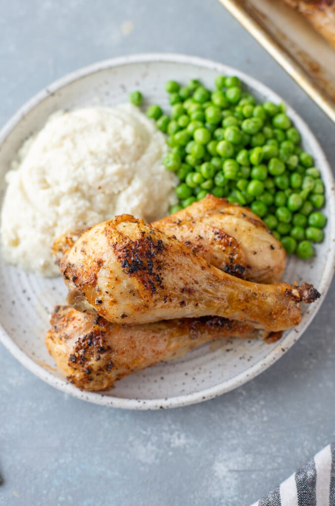 An overhead shot of 3 Baked Chicken Legs piled on a white plate with mashed potatoes and peas on the side on a grey background