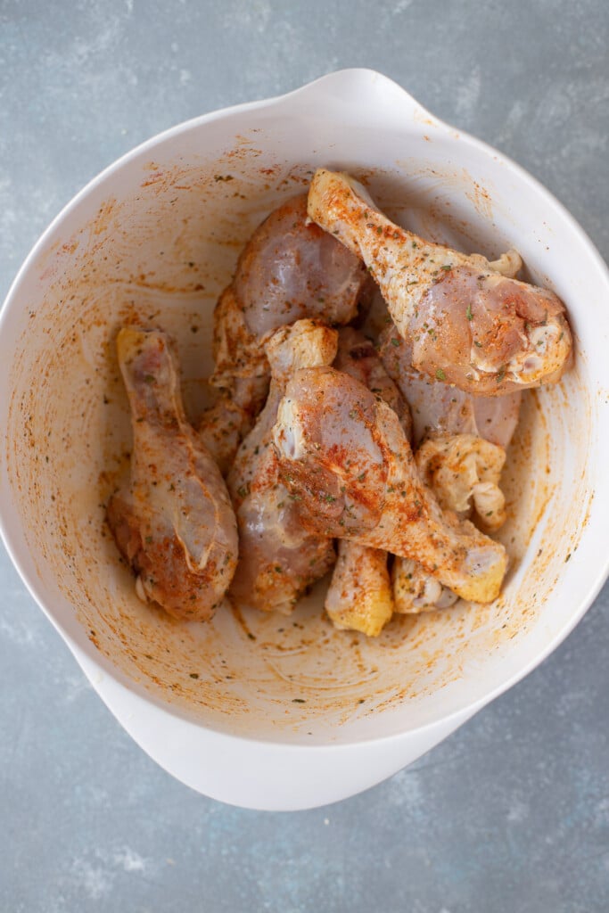An overhead shot of a white bowl with chicken drumsticks evenly coated in olive oil and spices
