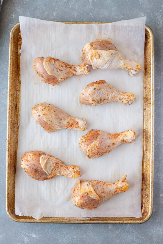 An overhead shot of 7 raw chicken drumsticks on a parchment lined baking sheet on a grey background