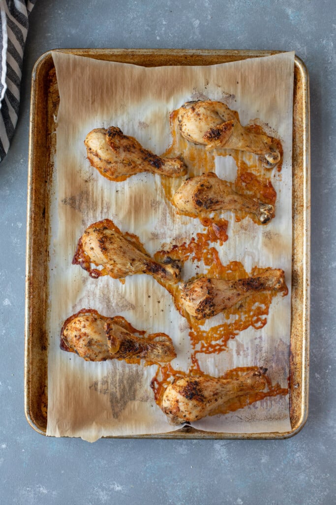 An overhead shot of 7 baked chicken legs on a parchment lined baking sheet on a grey background