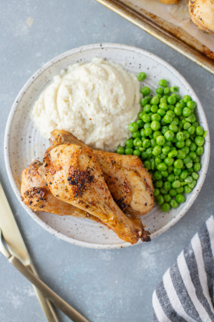 An overhead shot of 3 Baked Chicken Legs piled beside mashed potatoes and peas on a white plate with silverware and a baking sheet on the side on a grey background