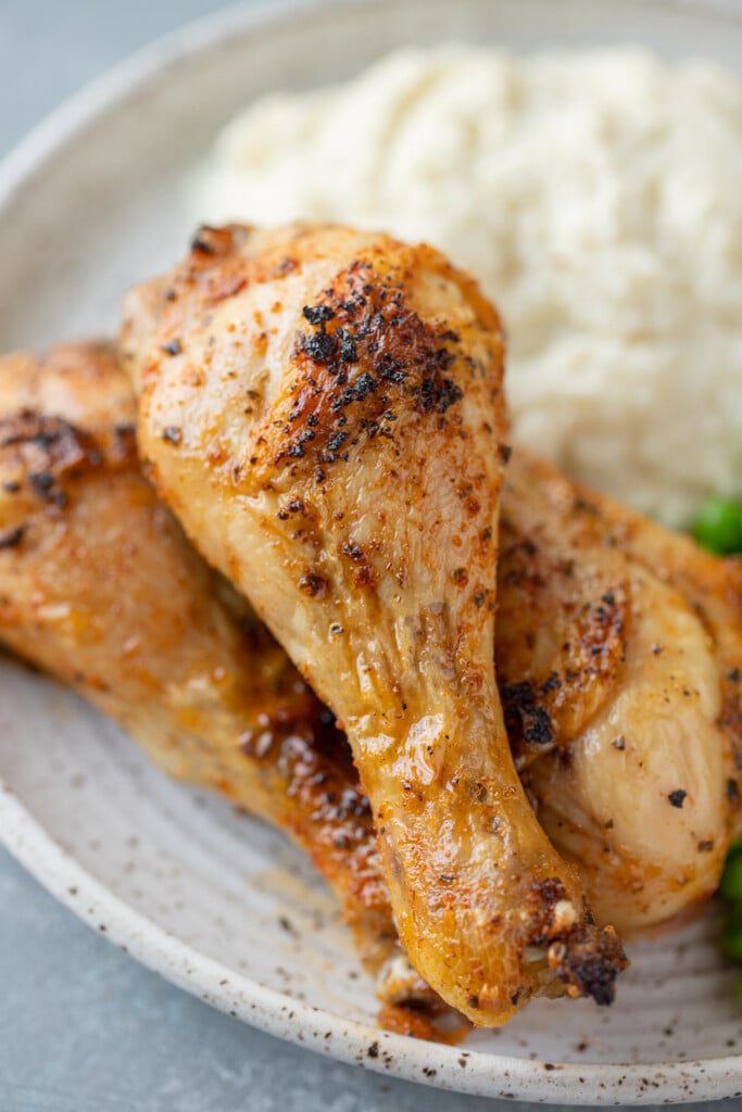A close up of 3 Baked Chicken Legs piled on a white plate with mashed potatoes on the side on a grey background