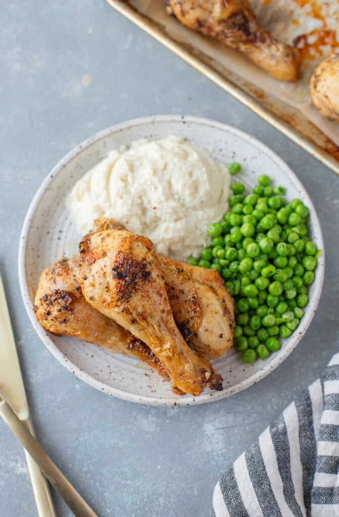 An overhead shot of 3 Baked Chicken Legs piled beside mashed potatoes and peas on a white plate with silverware and a baking sheet on the side on a grey background
