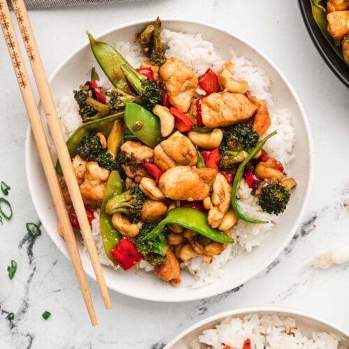 An overhead shot of a white plate with cashew chicken on a bed of white rice with chopsticks, another plate and a pan of cashew chicken on the side