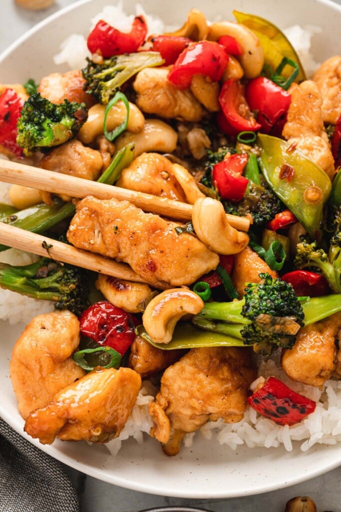 Cashew chicken on a bed of white rice on a white plate with chopsticks grabbing one piece of chicken