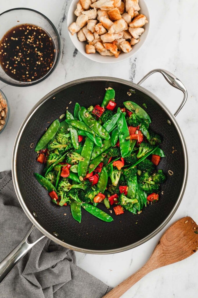 Broccoli, red bell pepper, snow peas, green onions, ginger, and garlic in a wok with chicken and cashew chicken sauce on the side