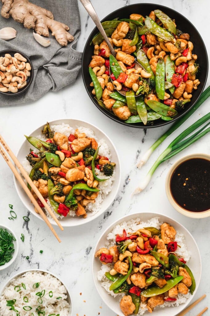 An overhead shot of 2 white plates with cashew chicken on a bed of white rice with chopsticks and a pan of cashew chicken on the side
