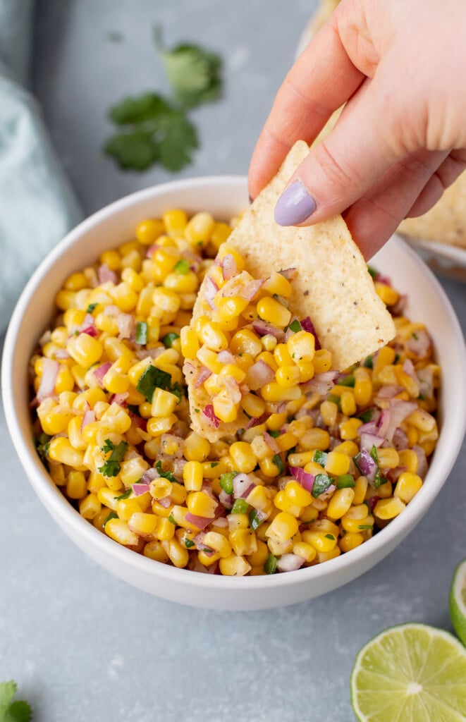 A white bowl of corn salsa with a hand holding a chip scooping out a large scoop of salsa on a grey background