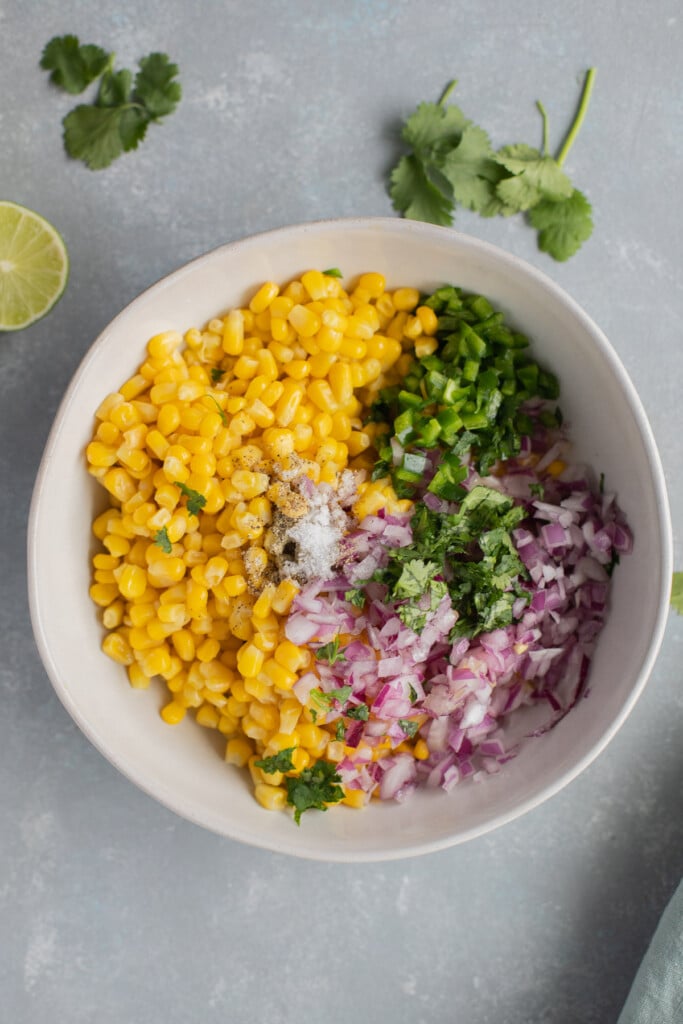 An overhead shot of a white bowl with the ingredients for corn salsa before being mixed on a grey background