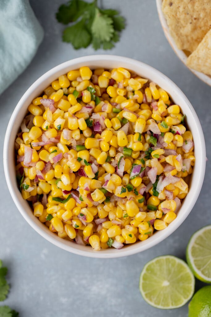 An overhead shot of a white bowl with corn salsa with a sliced lime, cilantro, and a bowl of chips on the side on a grey background