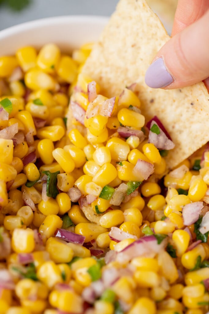 A hand holding a chip scooping out a large scoop of corn salsa from a white bowl