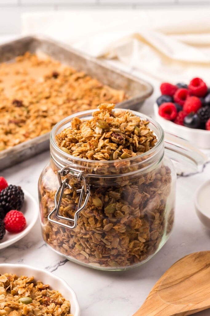 A clear small jar of homemade granola with a baking sheet of granola in the background and berries on the side