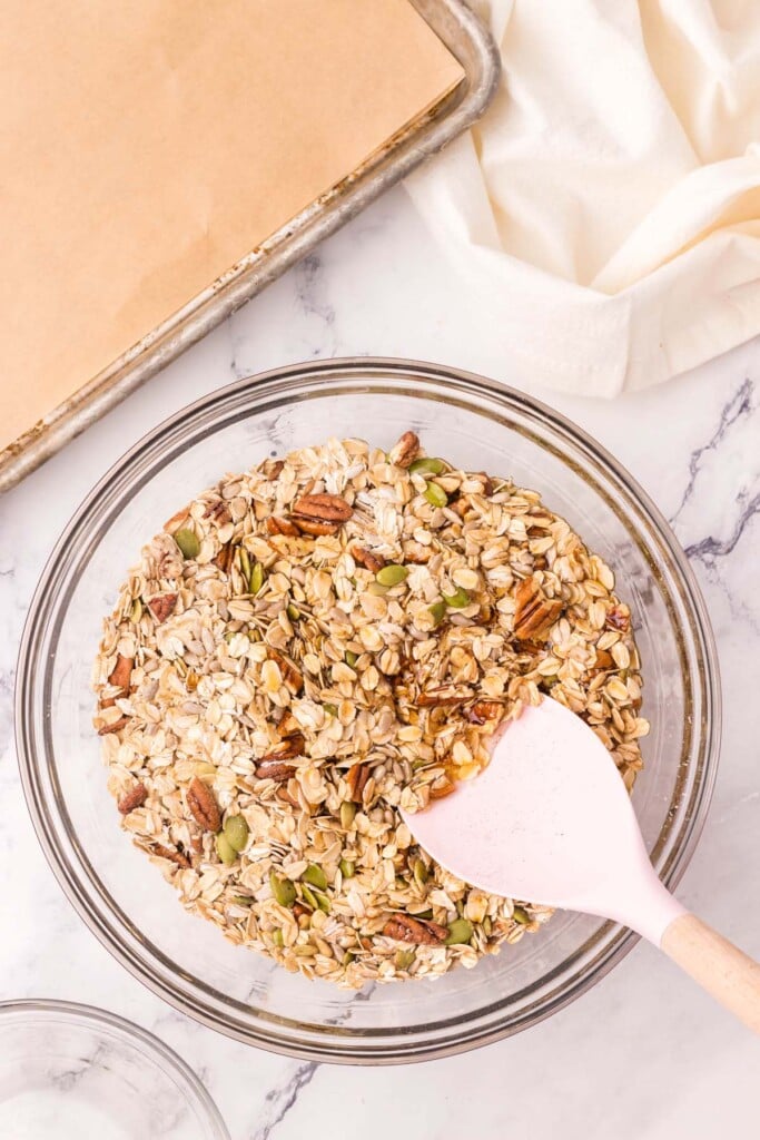 wet ingredients for homemade granola getting mixed into dry ingredients in a clear bowl with a spatula