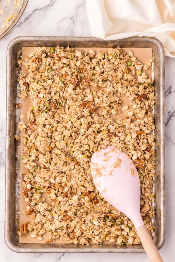 Uncooked homemade granola being spread with a spatula on a parchment lined baking sheet
