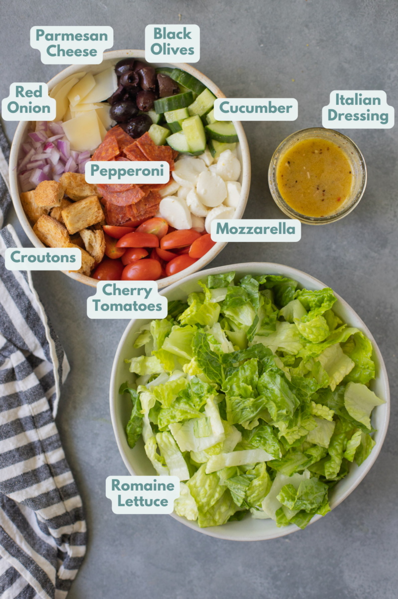 Ingredients for healthy italian salad in white bowls: romaine lettuce, pepperoni slices, cherry tomatoes, mini mozzarella balls, croutons, black olives, parmesan cheese, cucumber, red onion, italian dressing