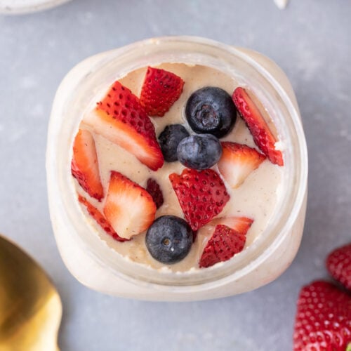 An overhead shot of protein overnight oats with strawberries and blueberries on top with a gold spoon on the side with a grey background