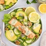 An overhead shot of a white bowl with salmon salad in it with avocado and lemon slices on it and another salad and tahini dressing on the side