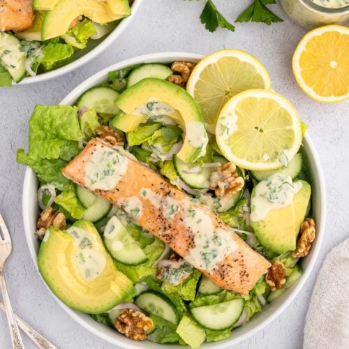An overhead shot of a white bowl with salmon salad in it with avocado and lemon slices on it and another salad and tahini dressing on the side