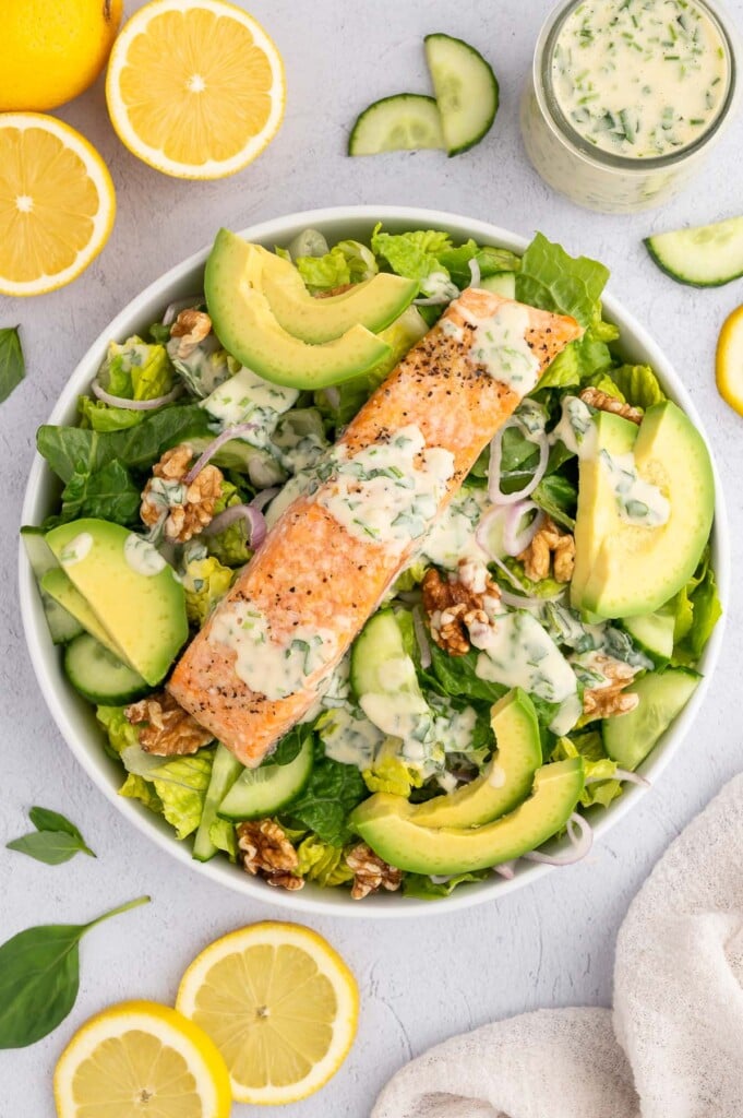 An overhead shot of a white bowl with salmon salad in it with avocado and lemon slices on it and tahini dressing and lemons on the side
