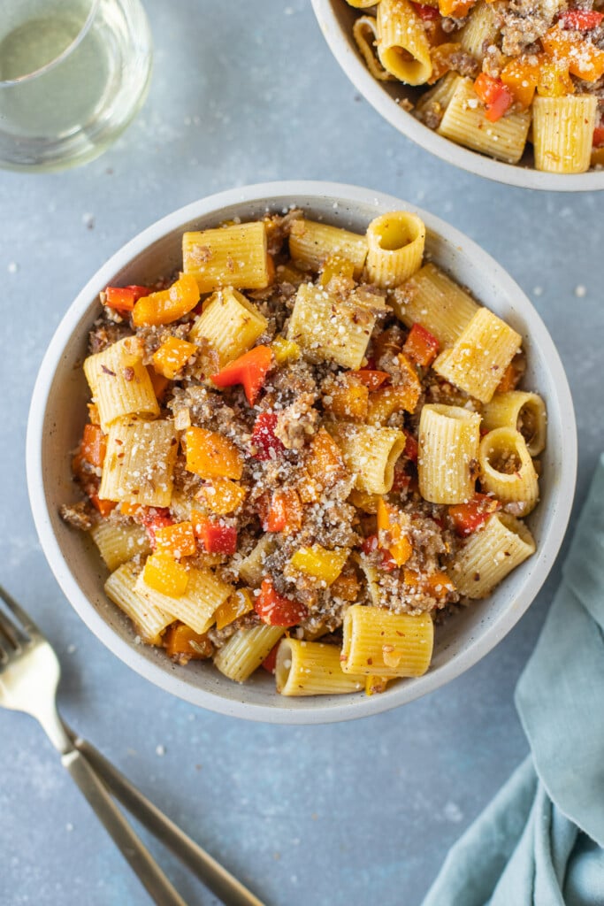 An overhead shot of a bowl of sausage and peppers pasta with forks and another bowl on the side on a grey background