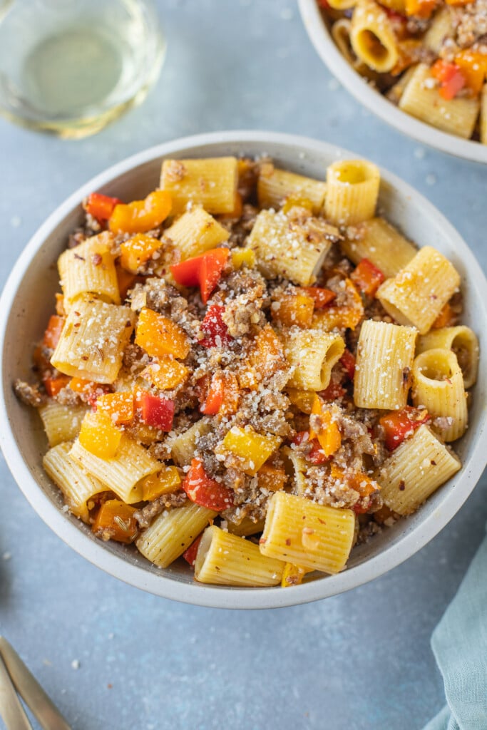 A bowl of sausage and peppers pasta on a grey background with another bowl and glass of white wine in the background