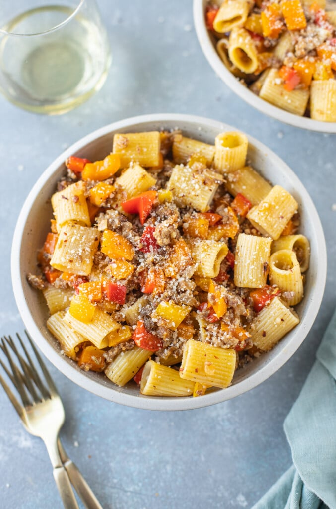 An overhead shot of a bowl of sausage and peppers pasta with forks, a glass of white wine, and another bowl on the side on a grey background