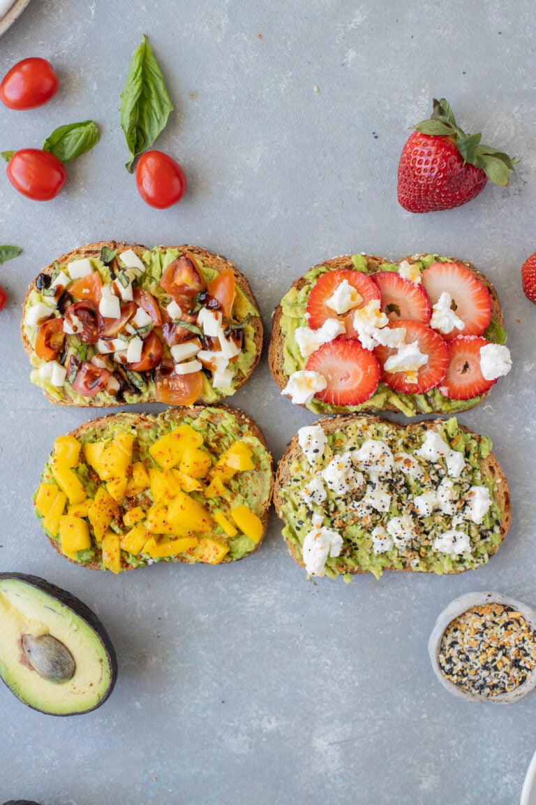 4 pieces of avocado toast (caprese toast, strawberry toast, mango tajin toast, goat cheese toast) on a grey background with tomatoes, basil, strawberries, avocado, and everything bagel seasoning on the side