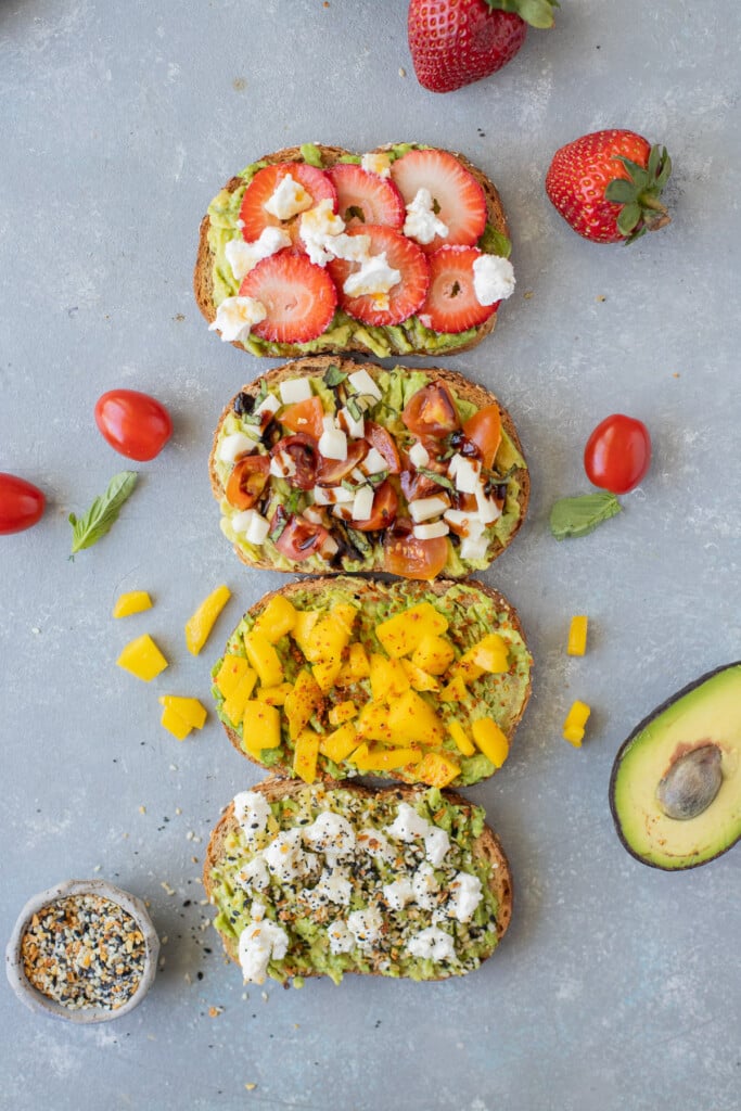 4 pieces of avocado toast in a row on a grey background (strawberry hot honey toast, caprese toast, mango tajin toast, goat cheese toast) with mango, avocado, everything bagel seasoning, tomatoes, adn strawberries on the side