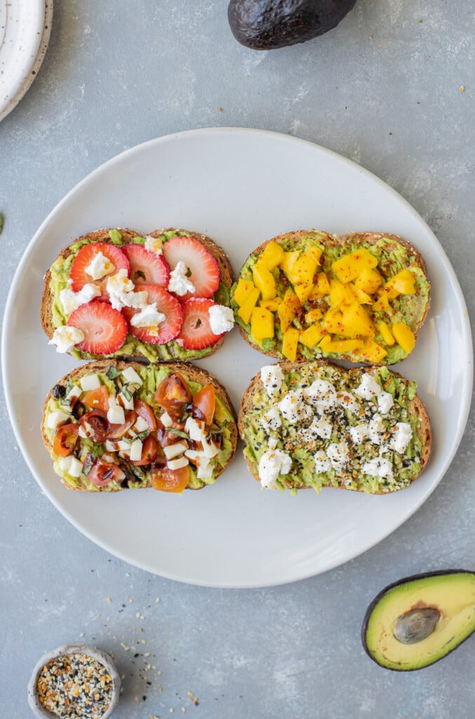 4 pieces of avocado toast (caprese toast, strawberry toast, mango tajin toast, goat cheese toast) on a white plate on a grey background with an avocado and everything bagel seasoning on the side