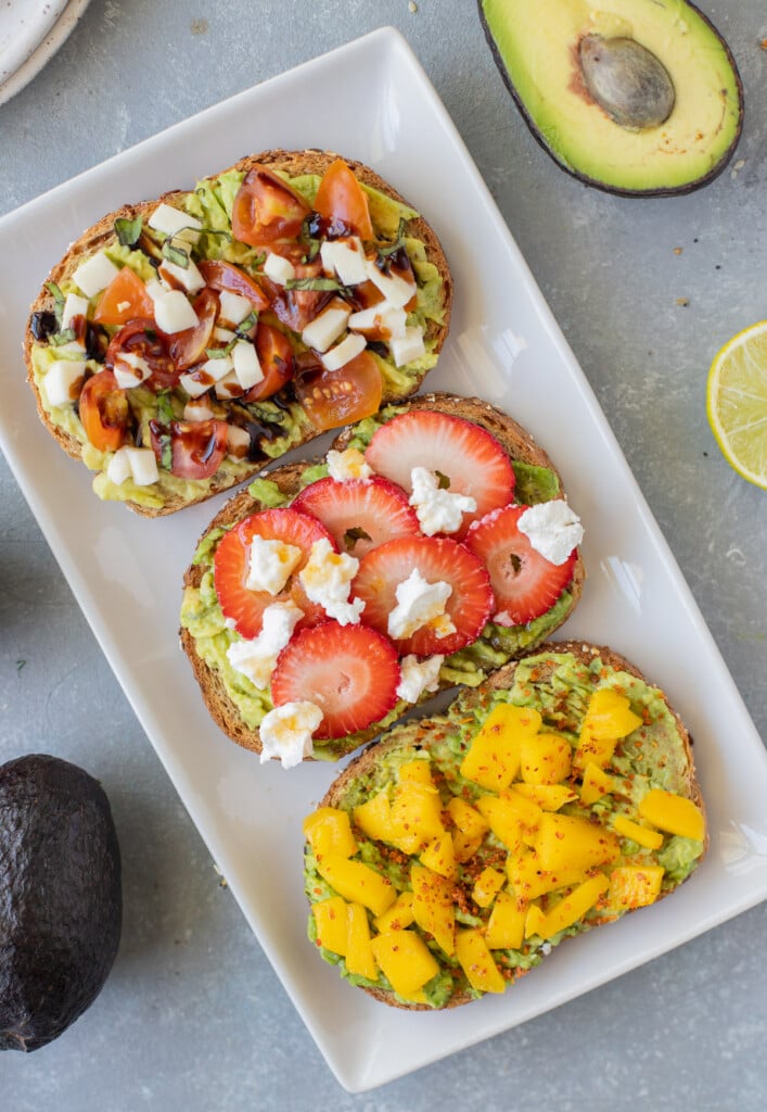 A white plate with 3 pieces of avocado toast on it (caprese toast, strawberry goat cheese, and mango tajin) with avocados on the side