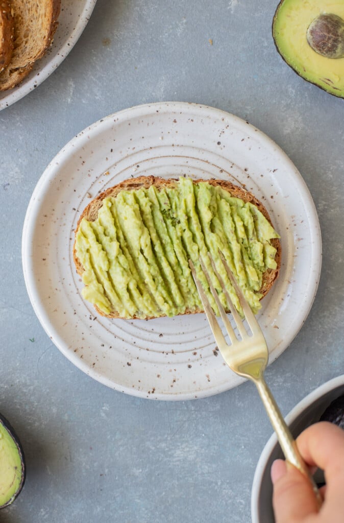 A piece of toast with a fork mashing avocado on it on a white plate on a grey background
