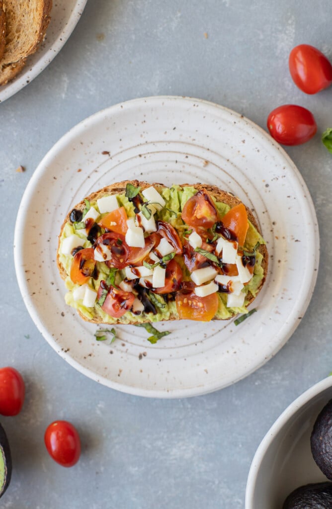 A piece of caprese avocado toast on a white plate with tomatoes on the side