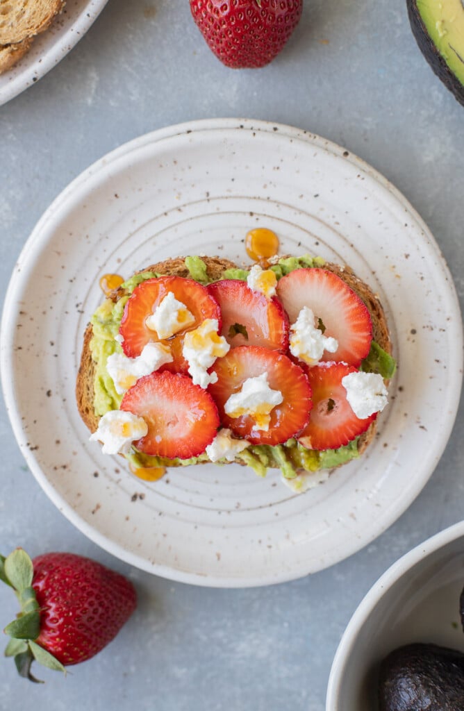 A piece of strawberry goat cheese avocado toast on a white plate with strawberries on the side