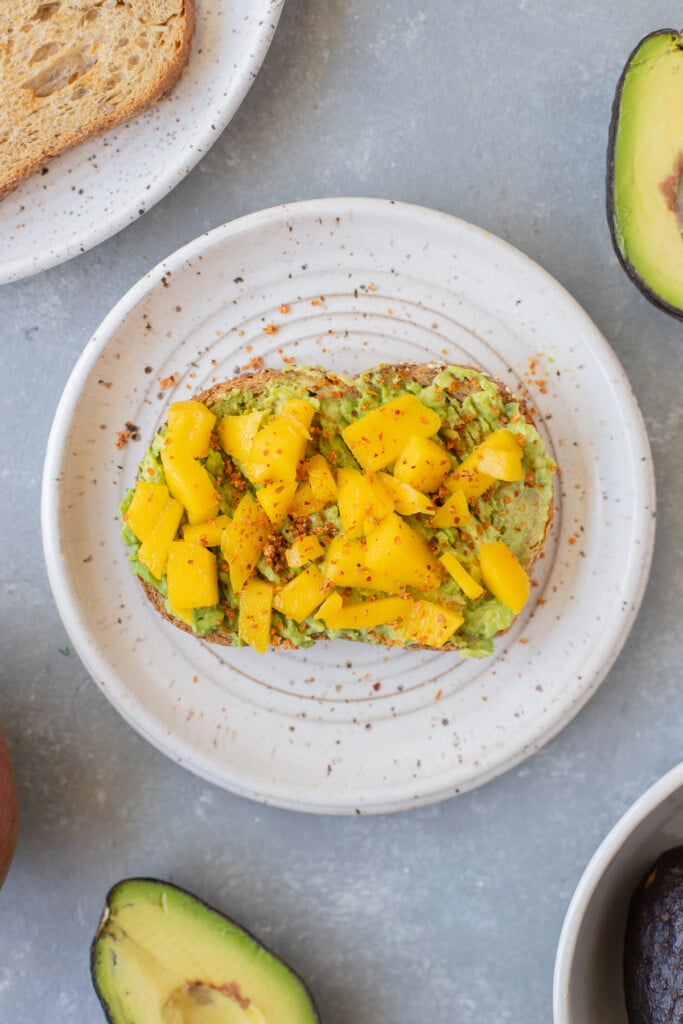 A piece of mango tajin avocado toast on a white plate with avocados on the side
