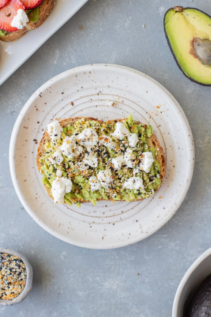 A piece of goat cheese avocado toast on a white plate with everything bagel seasoning on the side