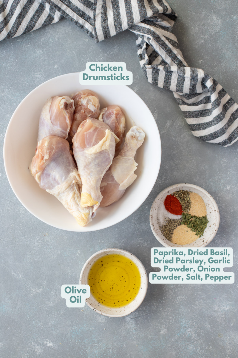 Ingredients for baked chicken legs in white bowls on a grey background: chicken drumsticks, olive oil, paprika, dried basil, dried parsley, garlic powder, onion powder, salt, and pepper