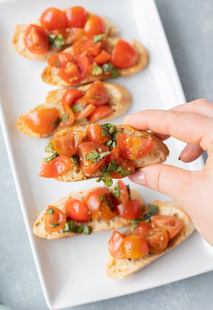 A hand holding up a bruschetta baguette toast with a white platter of more bruschetta toast in the background