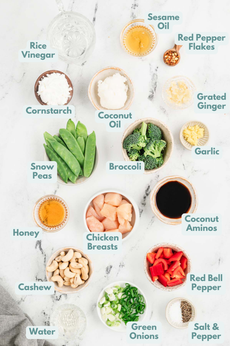 Ingredients for cashew chicken in small bowls on a white background: chicken breasts, salt, pepper, cornstarch, coconut oil, coconut oil, cashews, broccoli, red pepper, snow peas, green onions, ginger, garlic, coconut aminos, rice vinegar, water, honey, sesame oil, red pepper flakes