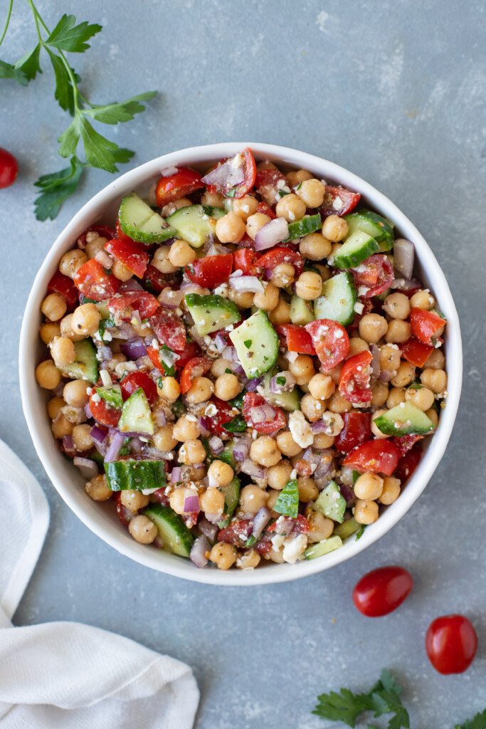 An overhead shot of a white bowl of Mediterranean chickpea salad on a grey background with tomatoes and herbs on the side