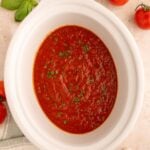 Healthy Crockpot Tomato Sauce in a white slow cooker base on a counter with tomatoes, herbs and garlic on the side.
