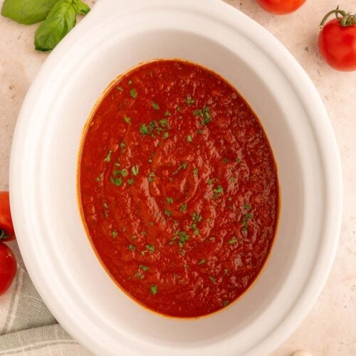 Healthy Crockpot Tomato Sauce in a white slow cooker base on a counter with tomatoes, herbs and garlic on the side.