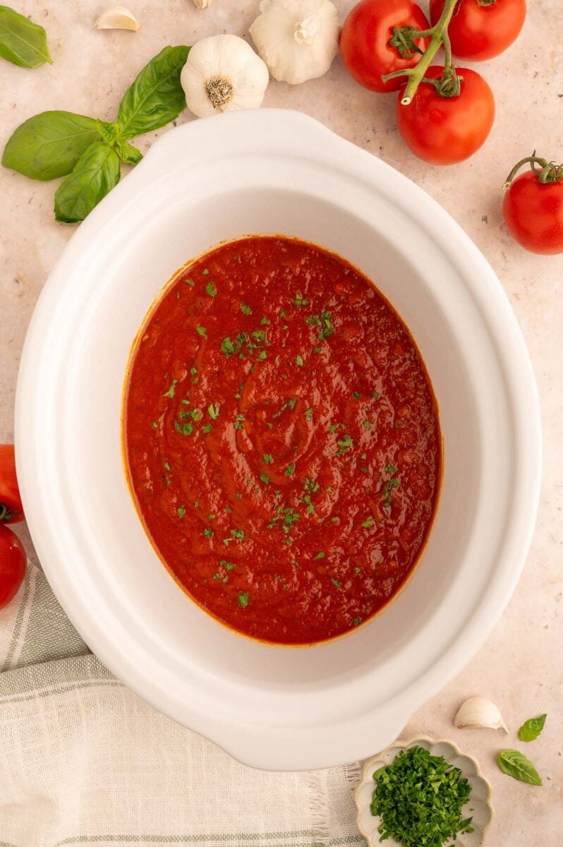 Healthy Crockpot Tomato Sauce in a white slow cooker base on a counter with tomatoes, herbs and garlic on the side.