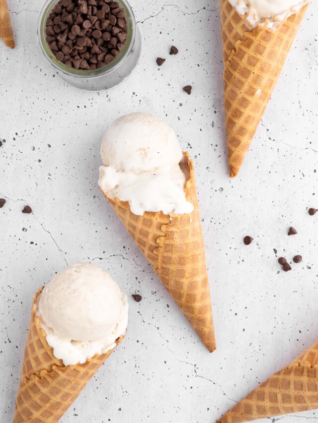 Vanilla protein ice cream in waffle cones on a white background with mini chocolate chips scattered around and a small bowl of them on the side