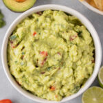 An overhead shot of the best guacamole in a white bowl with avocados, tomatoes, lime, and cilantro on the side