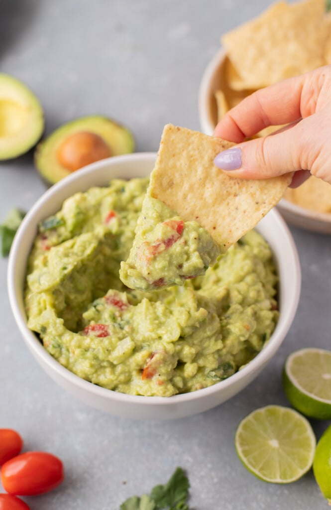 A tortilla chip taking a scoop out of a white bowl of guacamole with avocados, a bowl of chips, limes, tomatoes, and cilantro on the side