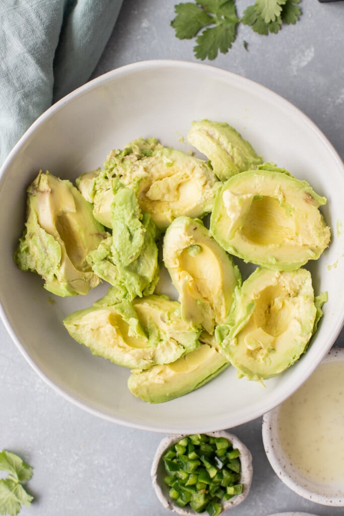 Avocados peeled and pitted in a white bowl before being mixed and mashed with cilantro and lime juice on the side