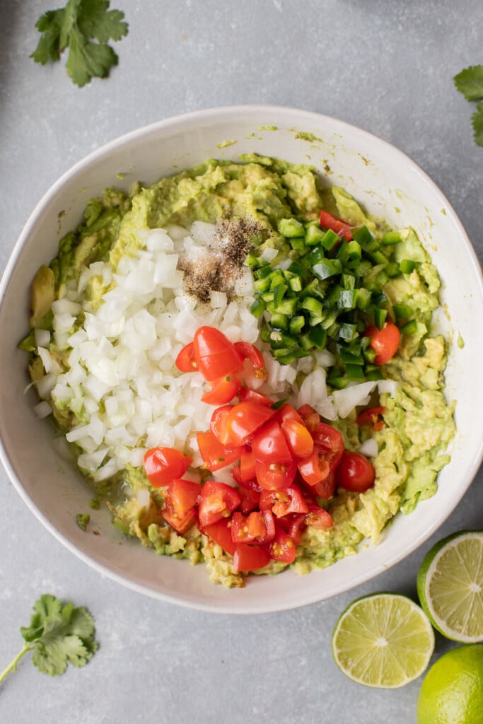 Ingredients for best guacamole in a large white bowl before being mixed together on a grey background