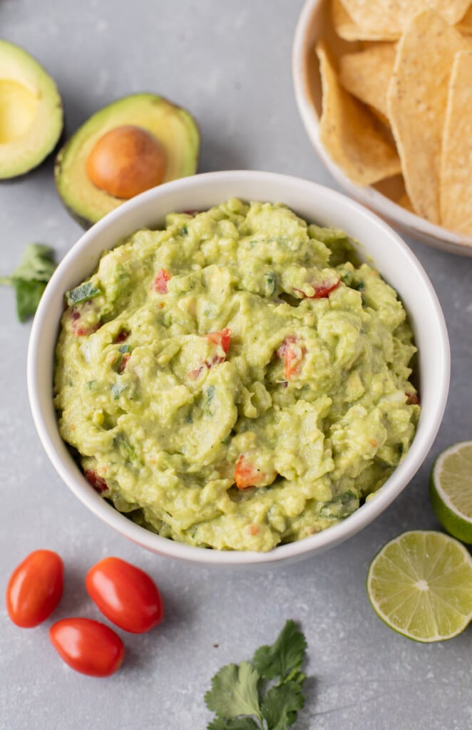the best guacamole in a white bowl with avocados, tomatoes, lime, a bowl of chips and cilantro on the side